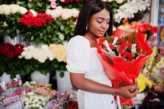 Hermosa chica afroamericana en tierno vestido blanco con ramo de flores en las manos de pie contra el fondo floral en la florería