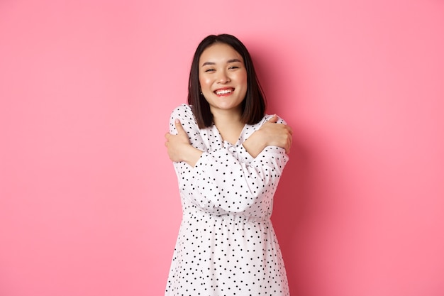 Hermosa chica asiática abrazando su propio cuerpo, abrazándose a sí misma y sonriendo con cara feliz, mirando a la cámara sin preocupaciones, de pie sobre fondo rosa