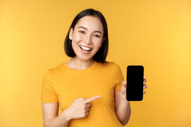 Hermosa chica asiática feliz que muestra la aplicación de pantalla del teléfono móvil en un dispositivo de teléfono inteligente de pie sobre fondo amarillo