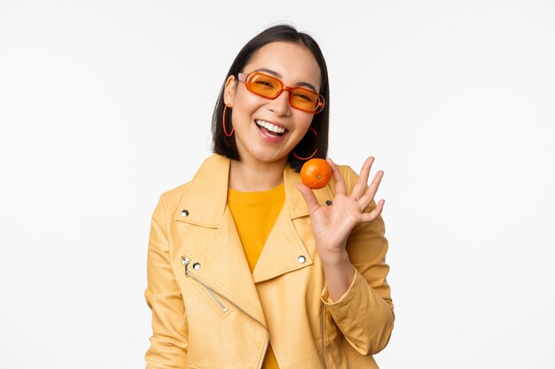 Hermosa chica asiática con gafas de sol mostrando mandarina y sonriendo feliz posando en amarillo contra el fondo del estudio