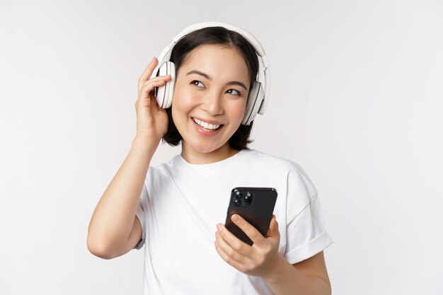 Hermosa chica asiática moderna escuchando música en auriculares con teléfono móvil usando la aplicación de teléfono inteligente de pie sobre fondo blanco