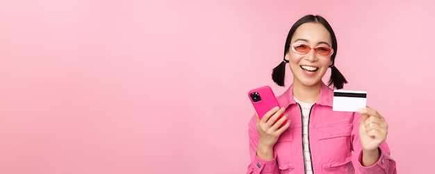 Hermosa chica asiática moderna riendo y sonriendo con tarjeta de crédito de teléfono móvil comprando en línea pagando con un teléfono inteligente de pie sobre fondo rosa