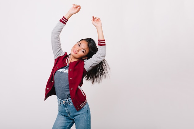 Hermosa chica asiática con piel ligeramente bronceada feliz bailando en la sala de luz. Adorable modelo de mujer en jeans con pelo negro lacio divirtiéndose frente a la pared blanca.