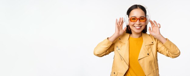 Hermosa chica asiática riendo feliz escuchando música en auriculares de pie sobre fondo blanco Copiar espacio