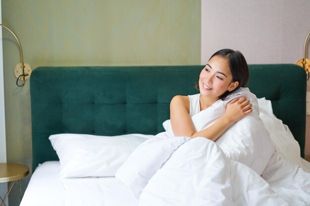 Hermosa chica asiática soñolienta se despierta en su cama con acogedoras mantas blancas mirando fuera de la ventana con