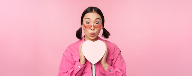 Hermosa chica asiática sonriendo feliz mostrando una caja de regalo de corazón y mirando emocionada a la cámara de pie sobre un fondo romántico rosa