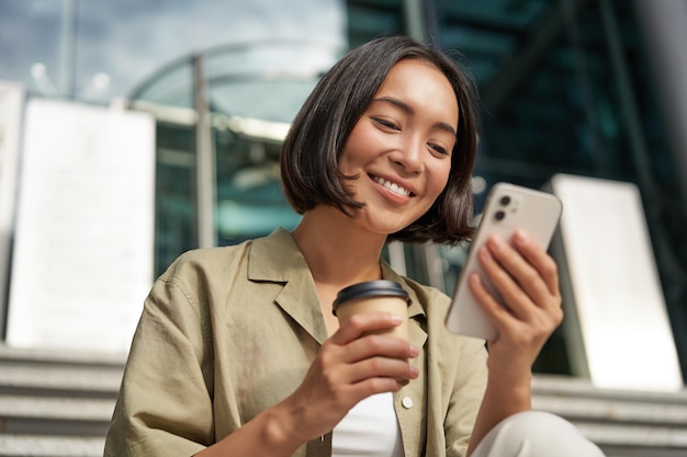 Hermosa chica asiática sonriente bebiendo café usando el teléfono móvil y sentada en las escaleras fuera de la joven mujer