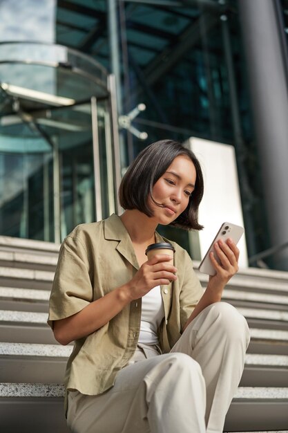 Hermosa chica asiática sonriente bebiendo café usando el teléfono móvil y sentada en las escaleras fuera de la joven mujer