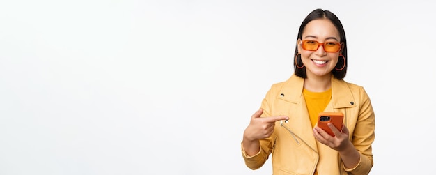 Hermosa chica asiática sonriente con gafas de sol apuntando con el dedo al teléfono inteligente que muestra la tienda de aplicaciones en el teléfono móvil de pie sobre fondo blanco