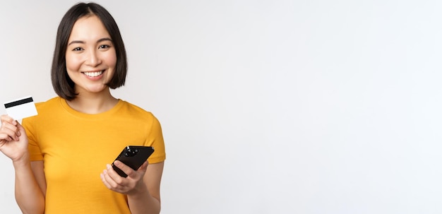 Hermosa chica asiática sonriente usando tarjeta de crédito y teléfono móvil pagando en línea en un teléfono inteligente de pie