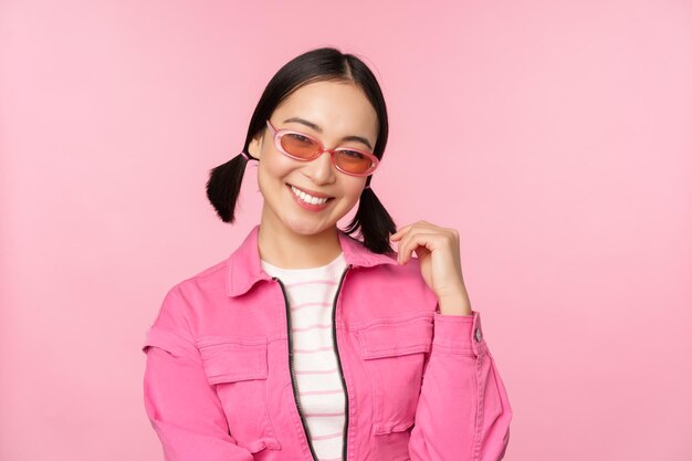 Hermosa chica asiática en sunlgasses sonriendo a la cámara posando sobre fondo de estudio rosa