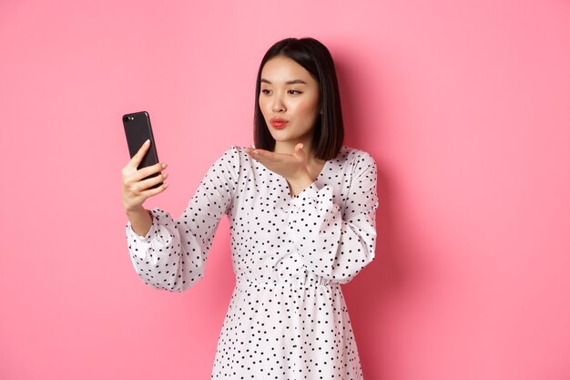 Hermosa chica asiática usando la aplicación de filtros de fotos y tomando selfie en el teléfono inteligente, posando con un lindo vestido sobre fondo rosa