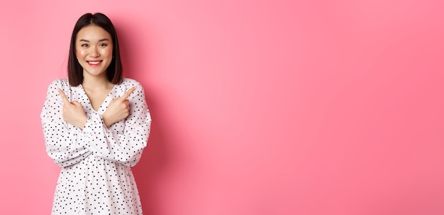 Foto gratuita hermosa chica asiática en vestido romántico apuntando con el dedo hacia los lados mostrando dos variantes en compras sm