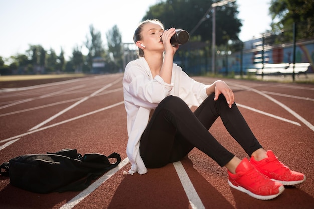 Hermosa chica en auriculares inalámbricos bebiendo agua pura mientras pasa tiempo en la pista de atletismo del estadio