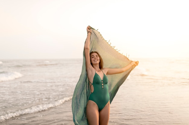 Hermosa chica en bikini verde con bufanda de pie cerca del mar