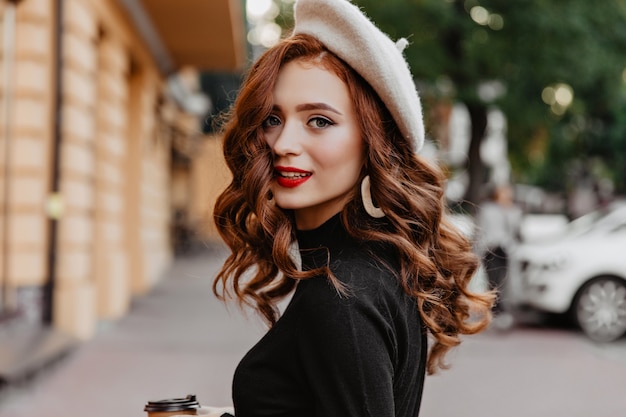 Hermosa chica blanca con cabello largo ondulado escalofriante en día de otoño. Retrato al aire libre del modelo femenino de jengibre interesado con taza de café.