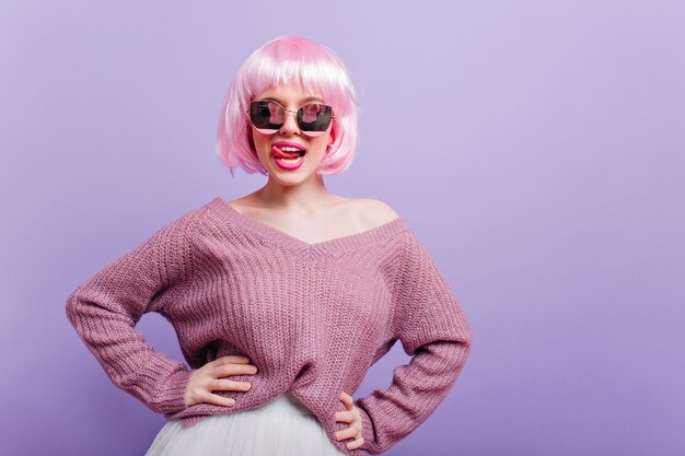 Hermosa chica blanca con peluca de moda divirtiéndose durante la sesión de fotos. Retrato de feliz modelo de mujer joven con pelo corto de color rosa de pie en pose de confianza y sonriendo.