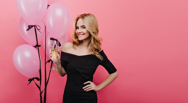 Hermosa chica blanca sosteniendo una copa de champán en la pared rosa. Bella dama con peinado ondulado disfrutando del vino mientras posa con globos de fiesta.