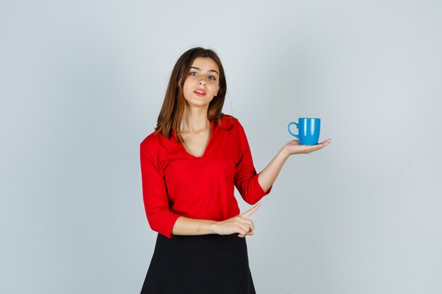 Hermosa chica en blusa roja, falda negra mostrando taza y mirando pensativo, vista frontal.