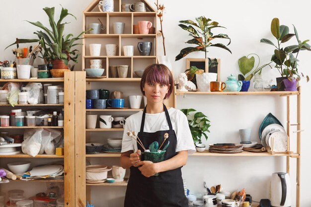 Hermosa chica con cabello colorido en delantal negro y camiseta blanca sosteniendo un tazón hecho a mano con una planta en las manos mirando soñadoramente en cámara al estudio de cerámica