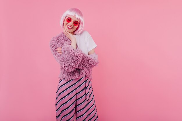 Hermosa chica con cabello corto rosa riendo con los ojos cerrados. jovencita refinada usa peruke y gafas de moda durante la sesión de fotos