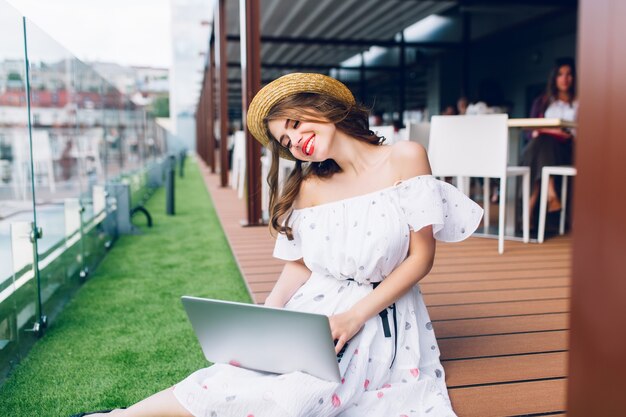 Hermosa chica con cabello largo está sentada en el piso de la terraza. Lleva un vestido blanco con hombros descubiertos, lápiz labial rojo y sombrero. Ella está escribiendo en la computadora portátil de rodillas.