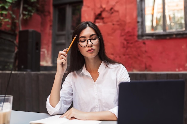 Hermosa chica con cabello oscuro en camisa blanca y anteojos trabajando cuidadosamente con una computadora portátil en el acogedor patio de la cafetería