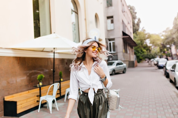 Foto gratuita hermosa chica con cabello rizado saludando caminando por la calle y mirando a su alrededor con una sonrisa