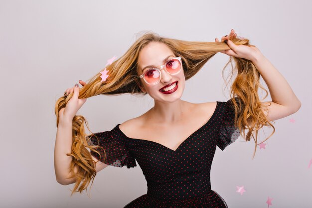 Hermosa chica con cabello rubio divirtiéndose, alegre joven en gafas rosa sonriendo. Tiene el pelo bastante largo y rizado en las manos, confeti de estrellas rosadas. Llevaba un bonito vestido negro. Aislado..