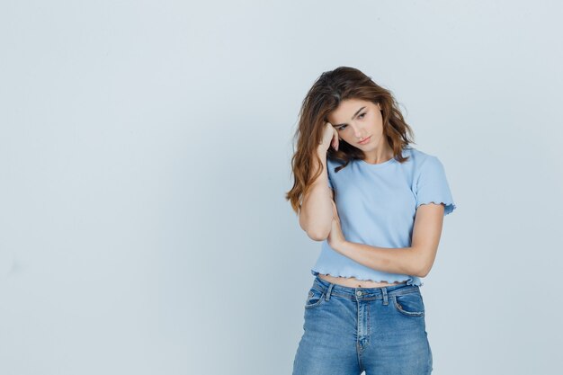 Hermosa chica con la cabeza inclinada en la mano en camiseta, jeans y mirando ansioso, vista frontal.