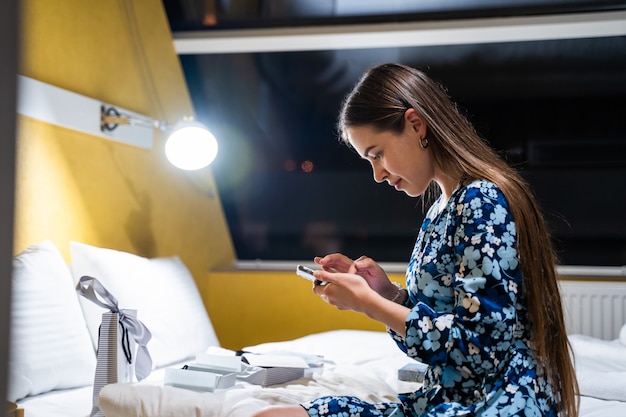 Hermosa chica en la cama en el dormitorio escribiendo en un teléfono inteligente móvil