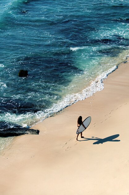 Hermosa chica camina con una tabla de surf en una playa salvaje. increíble vista desde la cima.