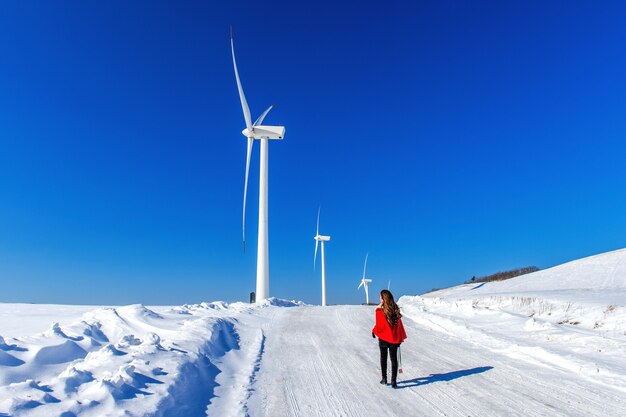 Hermosa chica caminando en el paisaje invernal del cielo y la carretera de invierno con nieve y vestido rojo y turbina eólica