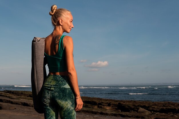 Hermosa chica caminando por la playa con estera de yoga