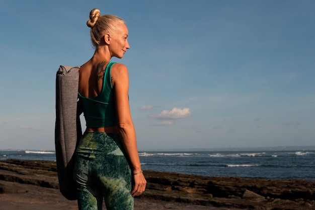 Hermosa chica caminando por la playa con estera de yoga