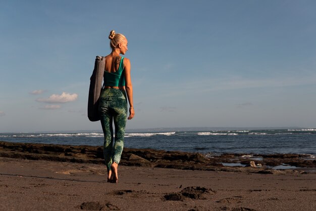 Hermosa chica caminando por la playa con estera de yoga