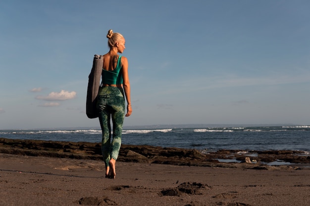 Hermosa chica caminando por la playa con estera de yoga