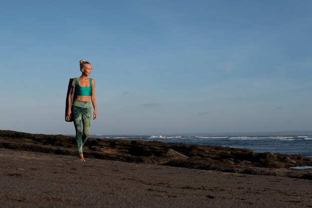 Hermosa chica caminando por la playa con estera de yoga