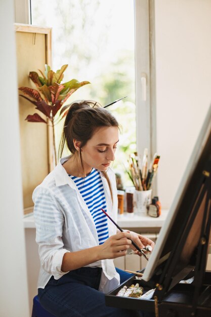 Hermosa chica con camisa blanca y camiseta a rayas preparándose soñadoramente para dibujar en caballete con ventana en el fondo en un hogar acogedor