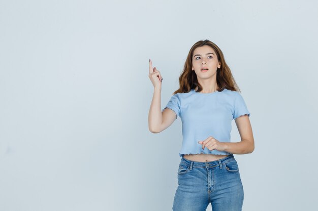 Hermosa chica en camiseta, jeans apuntando hacia arriba y mirando inteligente, vista frontal.