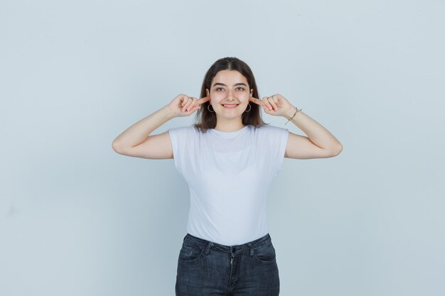 Hermosa chica en camiseta, jeans sosteniendo los dedos en las orejas y mirando alegre, vista frontal.