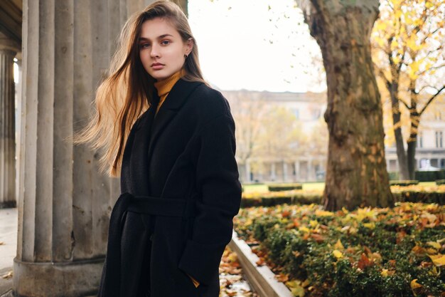 Hermosa chica casual con abrigo mirando con confianza a la cámara en el hermoso parque de la ciudad de otoño