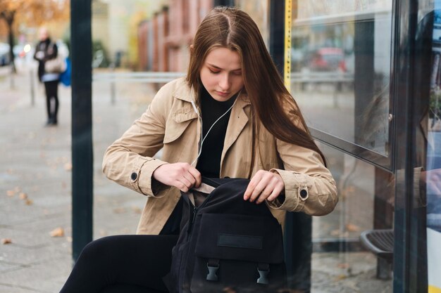 Hermosa chica casual en auriculares buscando algo en la mochila esperando el transporte público en la parada de autobús al aire libre