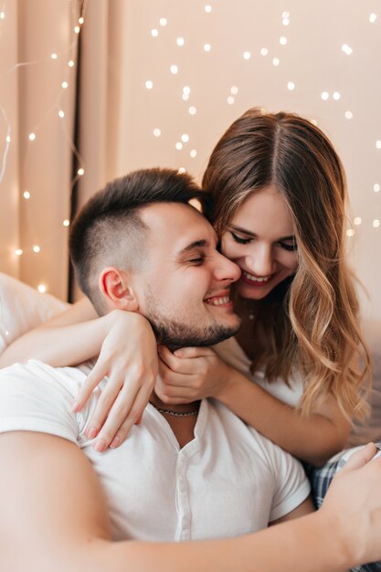 Hermosa chica caucásica abrazando suavemente a novio. Foto interior de dama riendo con cabello ondulado pasar tiempo con su marido morena.