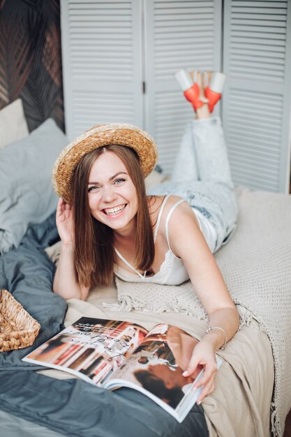 Hermosa chica caucásica con cabello oscuro y rizado, sombrero, camiseta blanca, jeans se encuentra en la gran habitación luminosa y lee una revista