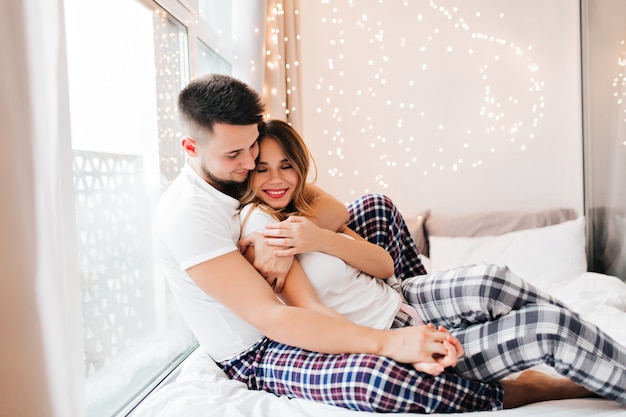 Hermosa chica caucásica cogidos de la mano con su novio mientras se relaja en casa. Foto interior de mujer refinada en pantalones a cuadros grises abrazando con su marido.