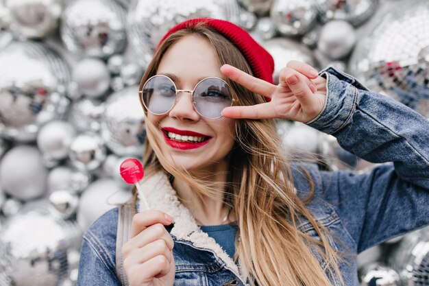 Hermosa chica caucásica comiendo dulces con sonrisa en la pared de brillo. Encantadora mujer rubia posando con piruleta cerca de bolas de discoteca.