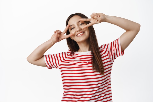 Hermosa chica caucásica que muestra signos de paz cerca de los ojos, sonriendo y luciendo feliz, con un estado de ánimo positivo de verano, parada en una camiseta roja contra el fondo blanco