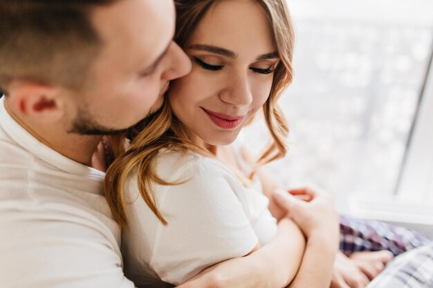 Hermosa chica caucásica sonriendo mientras su novio la abraza. Retrato interior de escalofriante modelo femenino abrazando con su marido en buenos días.