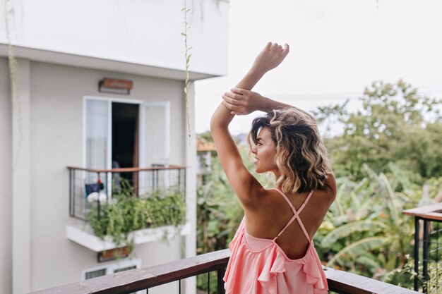 Hermosa chica caucásica en traje rosa que se extiende en el balcón del hotel. Magnífica mujer rizada disfrutando de vistas a la ciudad desde la terraza.